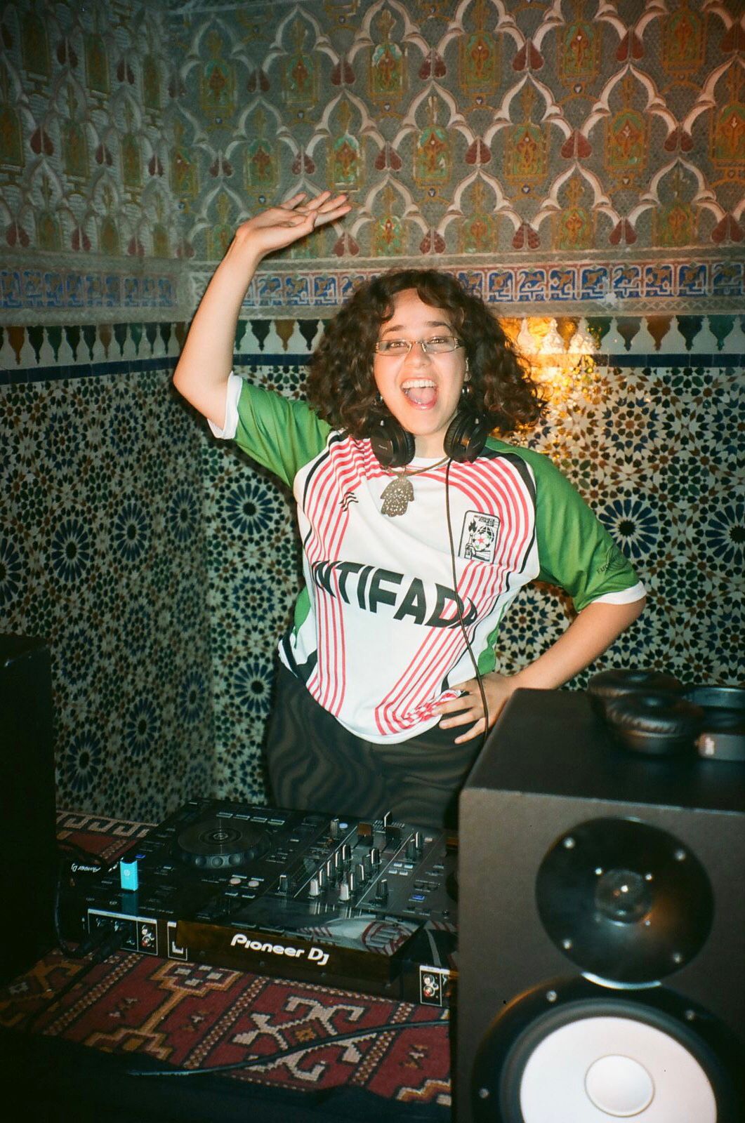 A photo of Balkis Guetta; she is smiling and waving at the camera while standing behind a turntable.